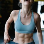 Fit young woman happily shows off weight-loss success by wearing oversized jeans in a gym.